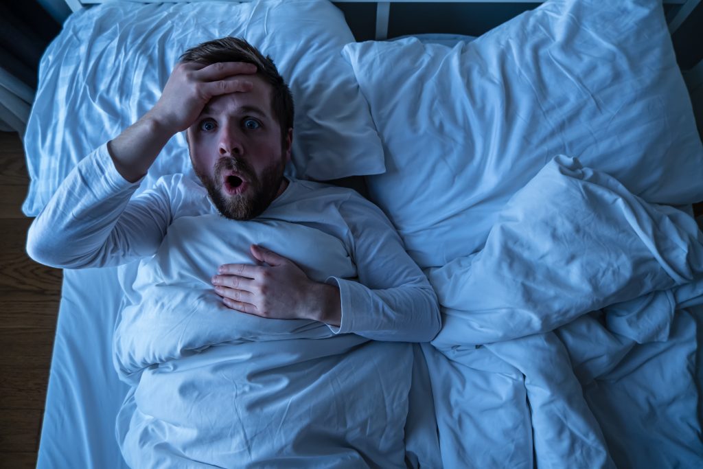 Mann im Bett mit erschrockenem Gesichtsausdruck, der einen Albtraum erlebt – Darstellung einer Parasomnie, die durch intensive Träume verursacht wird.