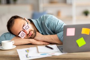 Mann schläft am Schreibtisch, trägt eine Brille mit Post-it-Zetteln, die Augen darstellen. Symbol für Müdigkeit im Büro, typisch für Schlafprobleme und Schlafmangel. Ein Schlaflabor wie SleepLab kann bei der Diagnose solcher Probleme helfen.