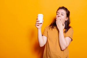 Frau in orangem T-Shirt, die gähnt und eine Tasse Kaffee hält – typisches Anzeichen von Müdigkeit und dem Bedürfnis nach einem Powernap.