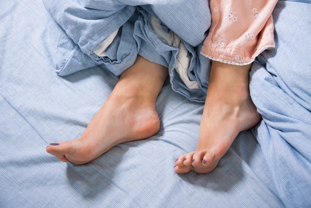 Eine Person im Bett, deren Beine aufgrund des Restless-Legs-Syndroms aus der Decke hervorschauen.