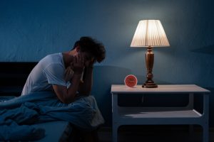 Mann sitzt schlaflos nachts auf seinem Bett, mit sichtbarem Stress, während eine Lampe schwaches Licht spendet. Schlafstörungen und nächtliche Schlafprobleme führen oft zu Schlafmangel und damit zu einem gestörten Schlaf-Wach-Rhythmus.