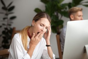 rau am Arbeitsplatz, die gähnt und sich an den Kopf fasst, aufgrund von Erschöpfung durch Schlafmangel. Müdigkeit und reduzierte Leistungsfähigkeit im Büro sind typische Auswirkungen von Schlafproblemen.