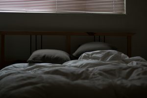 Ein Schlafzimmer mit einer schlichten, gemütlichen Einrichtung. Das Bett ist mit einer weichen, weißen Decke und zwei gepolsterten Kissen ausgestattet, die ein Gefühl von Ruhe und Entspannung vermitteln. Die Fensterjalousien sind geschlossen, wodurch das Zimmer dunkel und schlaffreundlich bleibt. Dieses Bild repräsentiert die perfekte Schlafumgebung, mit Fokus auf Dunkelheit, Ruhe und Komfort, die für die Erholung und Regeneration essenziell sind.
