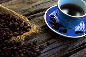 Nahaufnahme einer blauen Kaffeetasse mit frischem schwarzen Kaffee, umgeben von Kaffeebohnen auf einem rustikalen Holztisch. Symbolisiert den Einfluss von Koffein auf die Schlafqualität.