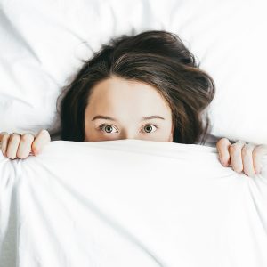 Eine Frau mit offenen, neugierigen Augen schaut unter einer weißen Bettdecke hervor. Das Bild symbolisiert den Übergang vom Schlaf zum Wachzustand.