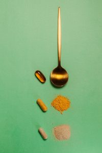 Ein goldener Löffel neben einer Auswahl an Nahrungsergänzungsmitteln, darunter Kapseln, Pulver und Tabletten, auf einem grünen Hintergrund. Das Bild symbolisiert die Vielfalt von Ashwagandha-Darreichungsformen.