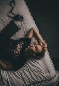 Eine Frau liegt entspannt auf einem Bett mit geschlossenen Augen und hört sanfte Einschlafmusik über ihre Kopfhörer. Neben ihr liegt eine Kamera, die eine entspannte und kreative Stimmung unterstreicht. Das sanfte Licht im Raum betont die abendliche Ruhe.