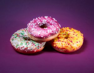 Drei bunt glasierte Donuts auf violettem Hintergrund stellen die Versuchungen ungesunder Ernährung dar. Dieses Bild illustriert die Herausforderung, die richtige Balance zwischen Genuss und Gesundheit zu finden.
