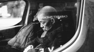 Ein älterer Mann schläft in sitzender Position in einem Fahrzeug, umgeben von reflektierenden Scheiben. Seine Haltung zeigt die Herausforderung von Schlaf in ungewohnten oder unbequemen Situationen. Das Bild thematisiert die Anpassung des Körpers an Schlaforte außerhalb des Bettes.