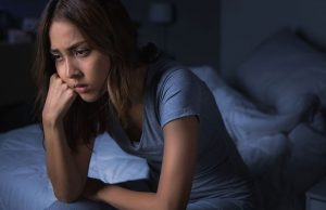 Eine Frau sitzt nachdenklich auf ihrem Bett in der Nacht – repräsentiert Schlaflosigkeit und deren Folgen wie Tagesmüdigkeit.