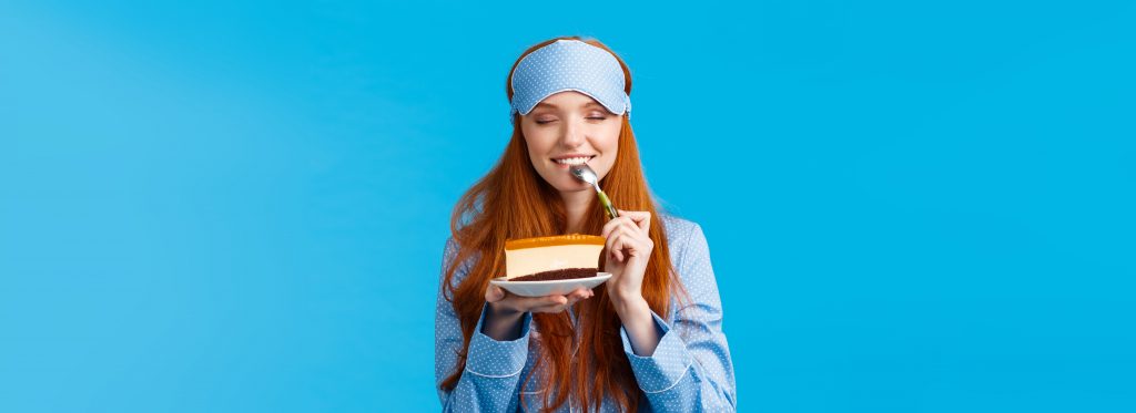 Eine junge Frau in Pyjama und Schlafmaske lächelt, während sie genüsslich ein Stück Kuchen isst. Die leuchtend blaue Hintergrundfarbe erzeugt eine fröhliche und entspannte Stimmung. Dieses Bild greift auf humorvolle Weise das Thema nächtliches Essen und seinen Einfluss auf den gesunden Schlaf in der Schweiz auf.