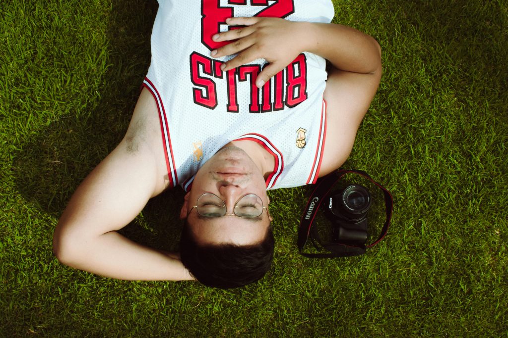 Ein junger Mann liegt entspannt auf einer grünen Wiese, trägt ein Basketballtrikot und hat die Augen geschlossen. Neben ihm liegt eine Kamera – ein Moment der Ruhe zwischen Sport und Fotografie. Solche Pausen sind essenziell für die Regeneration und damit auch ein Thema in der Schlaf Schweiz Beratung.